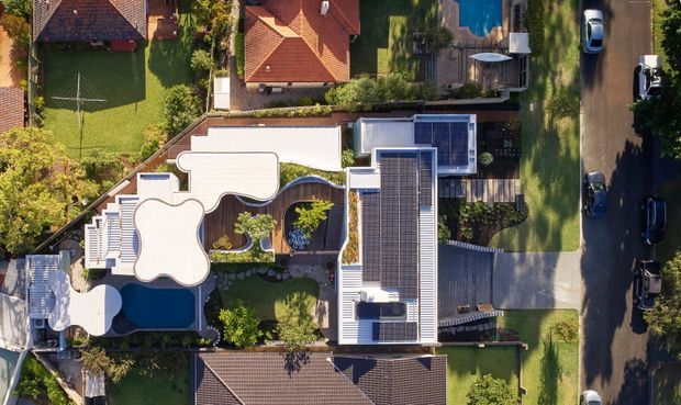 Aerial, top-down view of a contemporary residential property in a suburban neighbourhood. The house has a long rectilinear main structure with extensive rooftop solar panels, curved white roof forms, landscaped courtyards and garden areas, a swimming pool, and adjoining neighbouring homes and street parking.