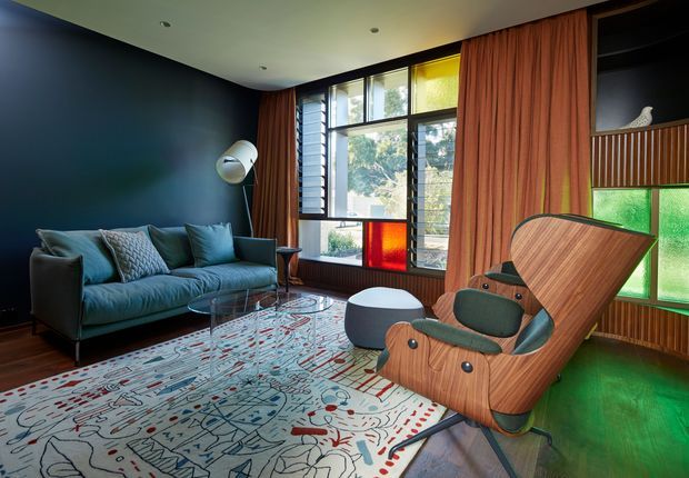 A richly styled mid-century-inspired living room with a deep navy feature wall, teal sofa, sculptural wooden lounge chair, patterned area rug, dark timber flooring, and dramatic coloured glazing. Full-height windows with timber framing and layered rust-orange curtains provide abundant natural light, while built-in timber panelled shelving and a green-tinted window add warmth and visual character.