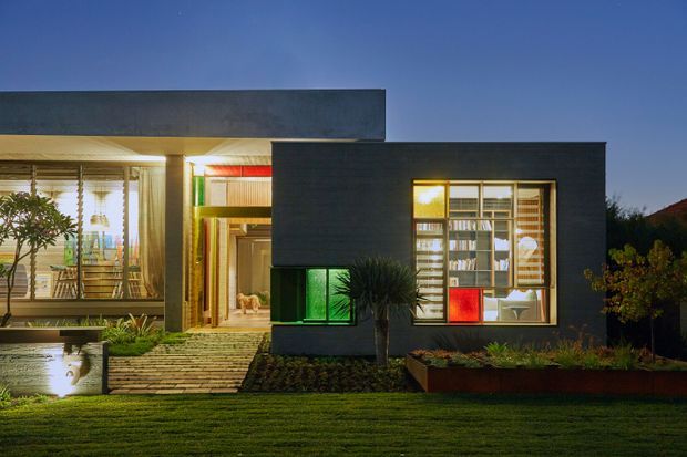 Twilight exterior view of a compact modern single-storey residence with strongly rectilinear flat-roofed volumes. Dark charcoal cladding contrasts with extensive warm-lit glazing, timber decking and a timber walkway leading through a landscaped front garden. Interior furnishings and shelving are visible through the windows, while sculptural planting and raised garden beds frame the facade.