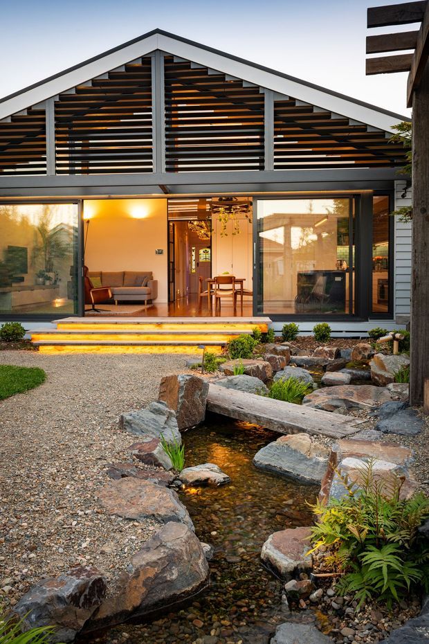 Exterior view of a contemporary gabled home with extensive glazing, a slatted timber screen beneath the roofline, and a warm illuminated living area opening onto a landscaped gravel garden. A narrow ornamental rock-lined stream runs through the foreground with stepping stones, planting, and a timber footbridge.