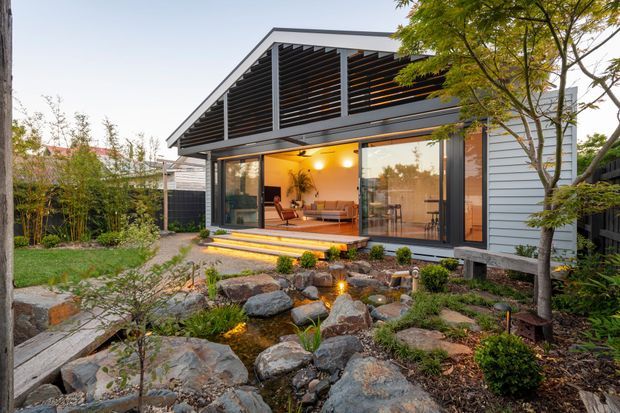 Exterior view of a contemporary single-storey pavilion-style home set within a landscaped garden. The building features grey horizontal cladding, extensive sliding glass doors, a dramatic slatted roof canopy, and a warmly lit open living area. A naturalistic rock garden with a small pond, stepping stones, and planted beds occupies the foreground.