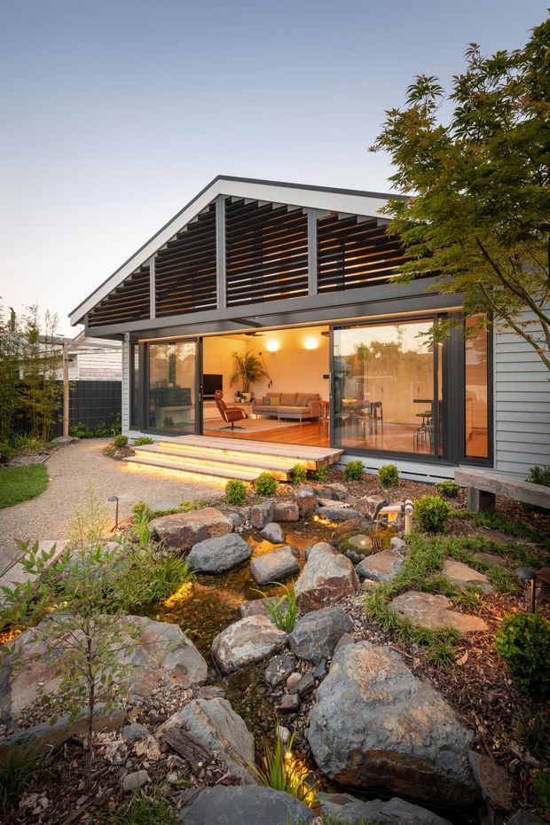 Exterior view of a contemporary single-storey house at dusk, featuring a steep gable roof, dark horizontal louvres, grey weatherboard cladding, extensive sliding glass doors, and a warm-lit open living area. A landscaped Japanese-inspired rock garden with a small stream and pond occupies the foreground.