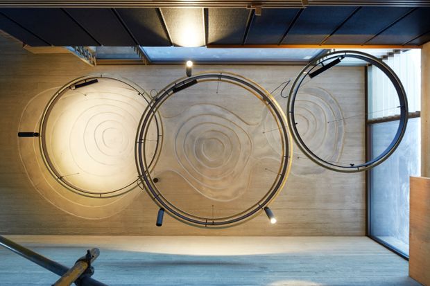 A modern interior viewed from above, featuring three large circular sculptural or track-like installations mounted against a warm concrete wall. The composition includes exposed structural and ceiling elements, linear dark ceiling panels, a glazed opening on the right, and a partially visible stair or railing in the foreground. The space has a striking geometric, industrial character with warm ambient lighting.