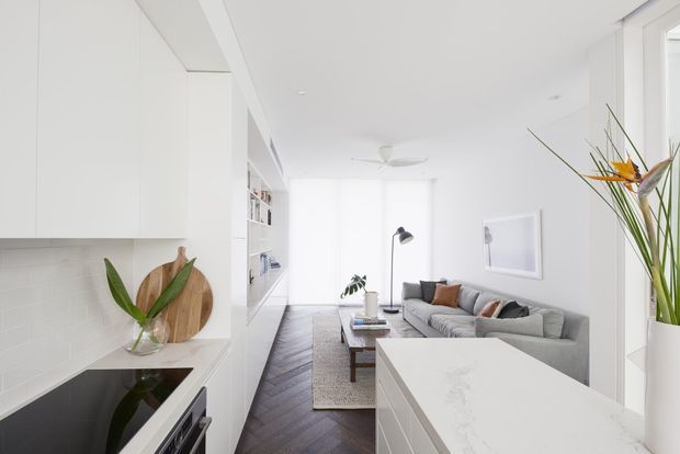 Bright open-plan interior showing a white contemporary kitchen in the foreground, with white cabinetry, a pale tiled splashback, dark timber herringbone flooring, and a marble-look island or benchtop. A grey sectional sofa and coffee table define the adjacent living area, with abundant white walls and daylight creating an airy feel.