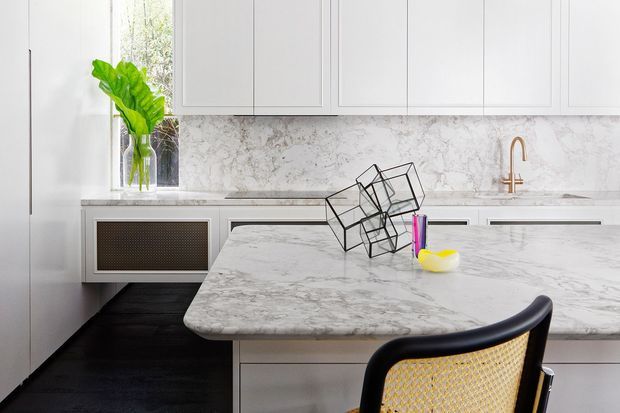 Bright contemporary kitchen featuring extensive white cabinetry, a veined marble-look backsplash and island benchtop, dark timber flooring, and a sculptural black-and-cane dining chair. A large island dominates the foreground, with a brass-toned tap and integrated appliances along the rear wall.