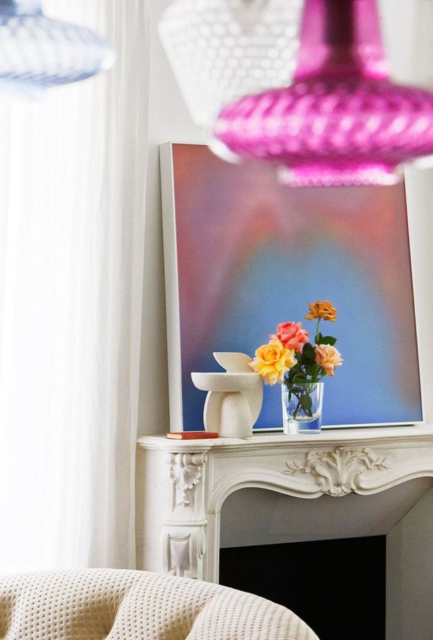 Bright, close-up interior vignette featuring an ornate white classical fireplace mantel with carved floral detailing, a colorful abstract artwork, a vase of flowers, a sculptural white object, and a cream tufted armchair. Sheer curtains and blurred pink pendant lighting frame the scene, creating a playful, eclectic atmosphere.