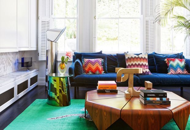 Colourful contemporary living room with a deep blue sectional sofa, vibrant patterned cushions, a sculptural timber coffee table, and a vivid turquoise rug. White built-in cabinetry, marble-look wall surfaces, tall shuttered windows, indoor plants, and eclectic decorative pieces create a bright, layered interior.