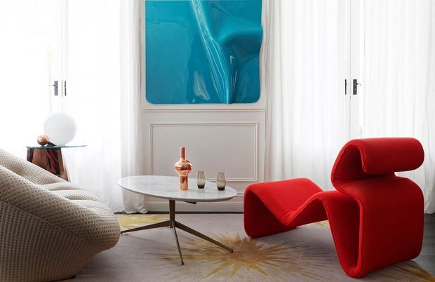 A refined contemporary living room with a sculptural bright-red lounge chair and matching angular ottoman beside a low oval coffee table. The room features white walls with decorative panel moulding, sheer white curtains, a large turquoise abstract artwork, pale flooring, a textured neutral sofa, and restrained metallic and glass accents.