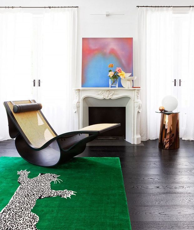 A bright, eclectic living room with tall sheer-curtained windows, dark timber flooring, and an ornate white fireplace mantel. A sculptural black lounge chair with woven cane panels sits on a vivid green rug featuring a white animal motif, while colourful abstract artwork and a small decorative side table add bold accents.