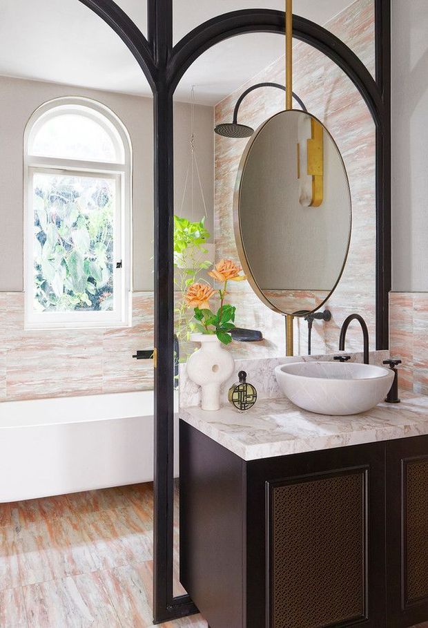 A refined bathroom featuring a freestanding white bathtub beneath an arched window, a dark framed glass shower partition, and a dark timber vanity with a white vessel basin. The vanity has a pale veined stone benchtop, large oval mirror, aged brass pendant and fittings, and black tapware. Pink and beige veined stone-look surfaces continue across the walls and floor, creating a warm, layered contemporary interior.