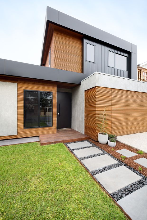 Exterior view of a contemporary multi-level residence with a strong geometric form. The facade combines dark grey vertical panel cladding, warm horizontal timber battens, smooth light rendered walls, and dark-framed glazing. A timber entry deck leads to a black front door, with a minimalist landscaped path of large concrete stepping slabs set in dark decorative stone beside a lawn.