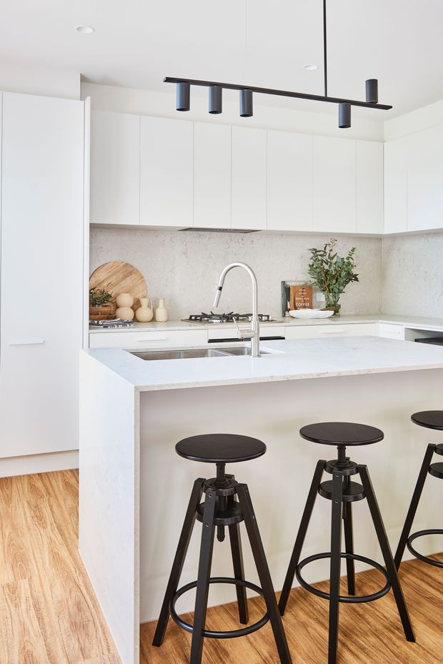 Bright contemporary kitchen featuring white full-height cabinetry, a pale stone benchtop and matching backsplash, an island with integrated sink and gooseneck tap, black linear pendant lighting, black industrial-style bar stools, and warm timber flooring.