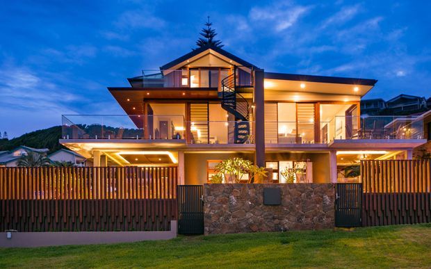Twilight exterior view of a contemporary multi-level residence with warm interior lighting, extensive glazing, projecting roof planes, balconies, and a prominent external spiral staircase. The property is enclosed by timber slat fencing and a natural stone boundary wall, with neighbouring hillside homes visible in the background.