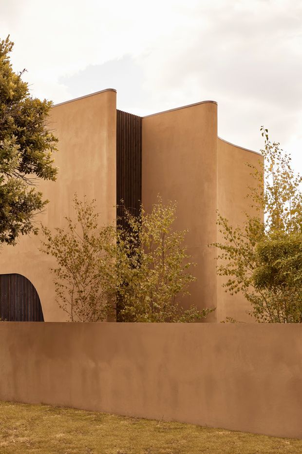 Exterior view of a sculptural contemporary building with warm terracotta rendered walls, curved parapet forms, a dark vertical slatted screen, and a rounded arched opening. The building is partially screened by mature landscaping and a matching rendered boundary wall, under an overcast sky.