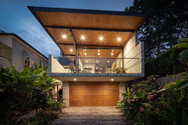 Twilight exterior view of a compact two-storey contemporary home set in lush landscaping. The upper level features a projecting flat-roofed balcony or living space with extensive glass walls, warm timber soffit panelling, recessed lighting, and a glass balustrade. Below is a wide timber-toned garage door framed by pale masonry, with a landscaped driveway and dense tropical planting in the foreground.