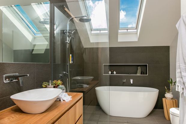 Contemporary bathroom with a freestanding white bathtub, vessel basin on a timber vanity, frameless glass shower, dark stone-look wall tiles, recessed shelf, and multiple skylights providing abundant natural light.