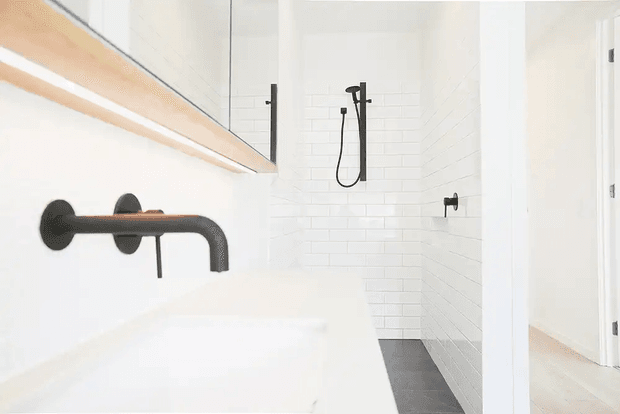 Bright, minimalist bathroom featuring a white tiled walk-in shower, matte black shower hardware and wall-mounted tap, a white vanity, and a large mirrored cabinet. A doorway opens to a light timber-floored adjoining space.