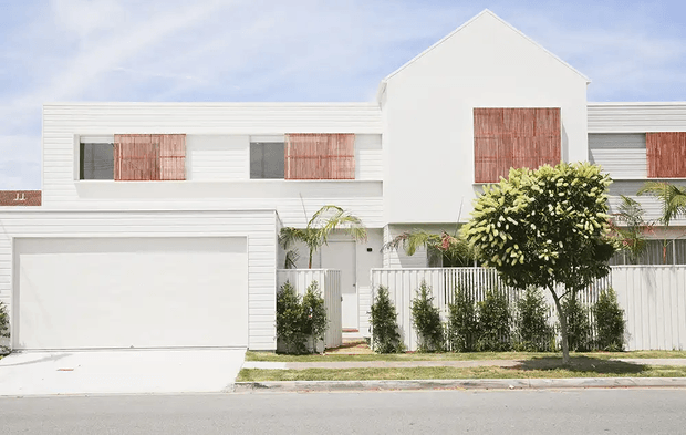 Front exterior view of a modern multi-storey townhouse with a crisp white rendered and horizontal-clad facade, prominent central gable, integrated double garage, upper-level windows with reddish timber shutters, and a fenced landscaped frontage.