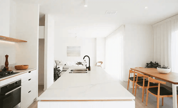 Bright, open-plan white kitchen and dining area. A large white island with integrated sink and dark gooseneck tap dominates the foreground, with white cabinetry, built-in oven and cooktop along the left wall. A timber dining table with matching timber chairs sits beside full-height sheer curtains, while light timber shelving and small natural-toned accents add warmth. The space is flooded with daylight and has a clean, minimalist appearance.