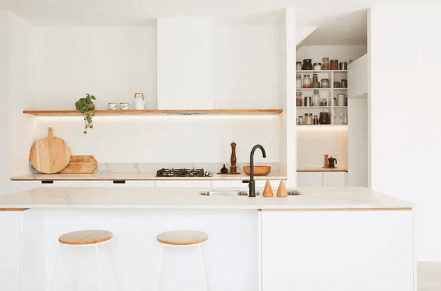 Bright minimalist white kitchen with a large island, integrated sink and curved black tap, gas cooktop, open shelving, and a walk-in pantry or storage recess displaying jars and kitchenware. Light timber shelving accents and warm wood chopping boards add contrast to the predominantly white cabinetry and surfaces.