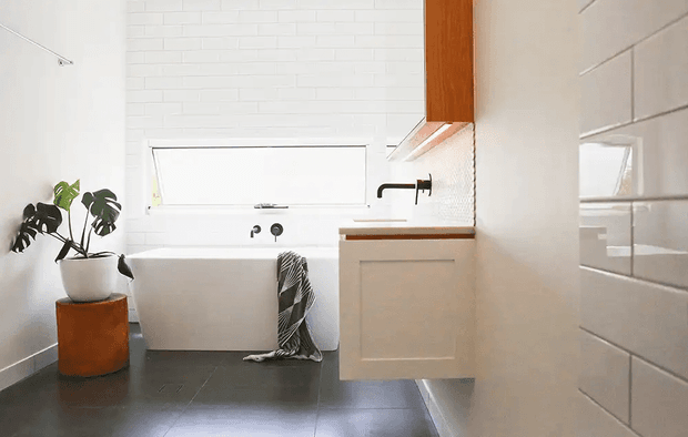 Bright contemporary bathroom with a freestanding white bathtub beneath a wide horizontal window, a wall-mounted timber vanity with dark benchtop, black tapware, white subway-tiled walls, and dark tiled flooring. A potted indoor plant and striped towel add warmth and contrast.
