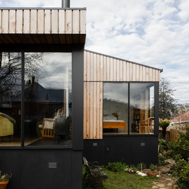 Exterior view of a contemporary residential extension with pale vertical timber cladding, dark metal lower walls and window frames, expansive floor-to-ceiling glazing, and low-pitched roof forms. The glazing reveals furnished interior rooms, while a small planted garden and paved path sit beside the building.