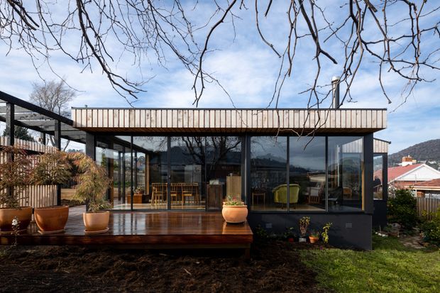 Exterior view of a contemporary single-storey home with extensive floor-to-ceiling glazing, dark window frames, and a projecting upper parapet clad in light vertical timber boards. A timber deck with potted plants fronts the glazed living area, while a covered outdoor structure extends to the left. Residential roofs and a wooded hillside are visible beyond the property.