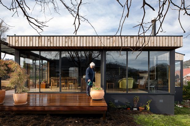 Contemporary single-storey pavilion-style house viewed from the garden, with extensive floor-to-ceiling glazing, dark framed windows, a low flat roof, and a warm timber deck. The transparent facade reveals a dining area and living spaces inside, while mature branches and landscaping frame the scene.