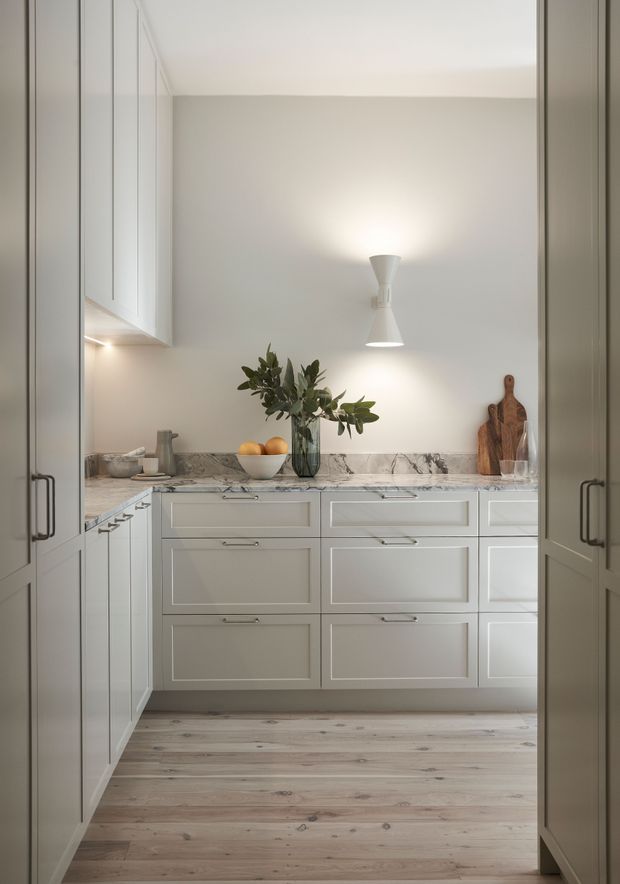 A compact, bright kitchen viewed through open tall cabinet doors. The room features pale grey-white shaker-style cabinetry with framed drawer fronts and slim metal handles, a grey-white veined stone benchtop, light timber floorboards, under-cabinet lighting, and a simple central wall light. A vase with greenery, fruit, and timber chopping boards provide restrained natural accents.