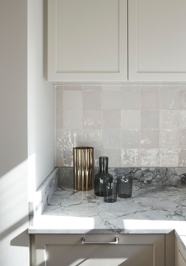 Close-up of a refined kitchen corner showing pale painted cabinetry, a glossy handmade-look tiled splashback, and a veined marble or natural-stone benchtop with matching upstand. Dark glass vessels and a vertically fluted metallic container sit on the counter, with strong sunlight creating diagonal shadows.