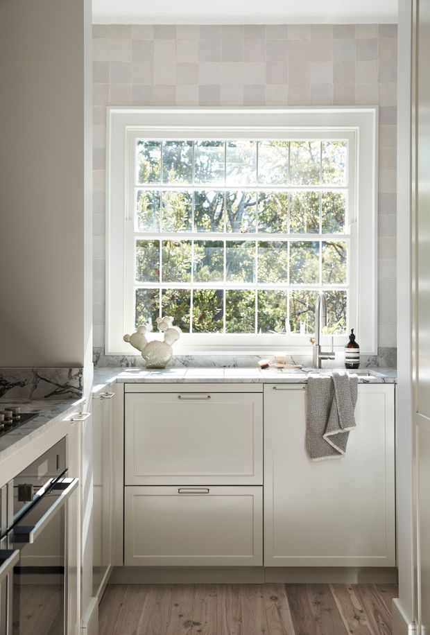 Bright, compact traditional kitchen with white Shaker-style cabinetry, a marble-look countertop, and a large multi-pane window above the sink. Soft pale square tiles form the backsplash, while timber flooring and brushed metal fixtures add warmth and contrast. The image includes a partial cooktop and built-in oven at left, with natural greenery visible through the window.