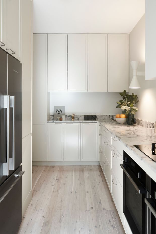 A bright, narrow galley kitchen with white shaker-style cabinetry, extensive white marble-look benchtops and splashback, integrated appliances, and pale timber flooring. A dark stainless-steel refrigerator occupies the left side, while black appliances contrast with the light cabinetry. Decorative pendant lighting, a vase of foliage, and a bowl of fruit add warmth.