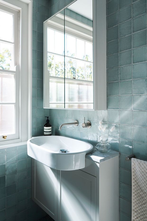 A bright, compact bathroom featuring a white wall-mounted basin with integrated vanity cabinetry, brushed metal wall-mounted tapware, a large mirrored cabinet, and glossy blue-green square wall tiles. White-framed windows provide abundant natural light, with a textured towel hanging at the side.
