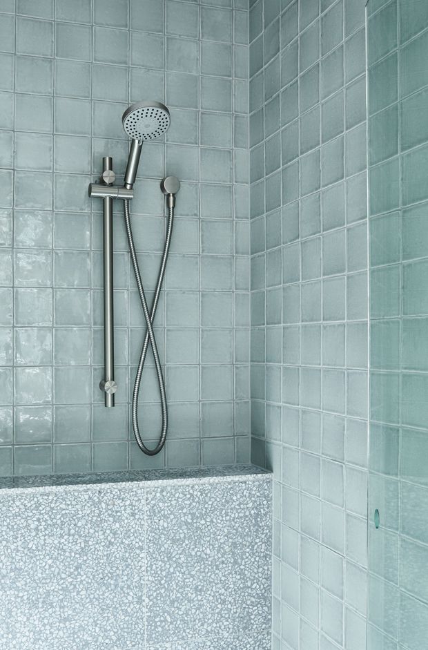 Close-up of a compact shower enclosure with pale blue-green square tiled walls, a brushed metal handheld shower rail and hose, and a terrazzo-finished partial partition or ledge in the foreground. The space is clean and contemporary with cool-toned finishes.