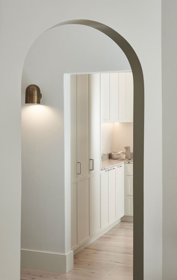 A softly lit interior view through a tall rounded archway into a compact white kitchen or pantry. The space features full-height panelled cabinetry, a pale stone benchtop, under-cabinet lighting, light timber flooring, white walls with traditional skirting, and a bronze wall sconce beside the opening.