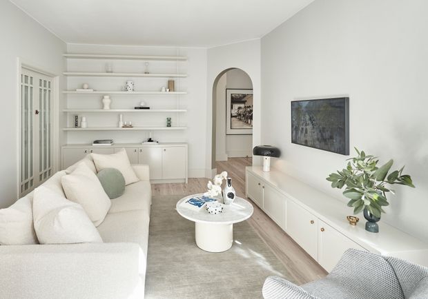 Bright, refined living room with a large cream sectional sofa, round white coffee table, pale timber-look flooring, and extensive white built-in shelving and cabinetry. A wall-mounted television, arched opening, framed artwork, soft neutral furnishings, and indoor plants create a calm contemporary interior.