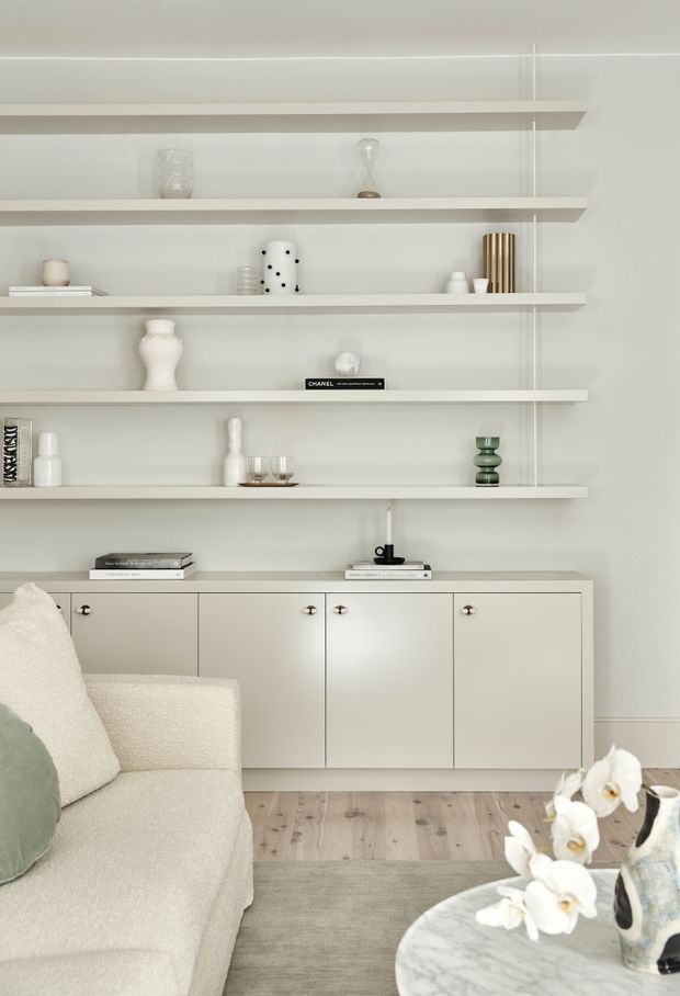 A serene, light-filled living room with minimalist built-in wall shelving and a low cream cabinetry unit displaying curated decorative objects and books. A softly textured cream sofa with a muted sage cushion sits beside a round marble coffee table, with pale timber flooring and neutral furnishings creating a refined, calm atmosphere.