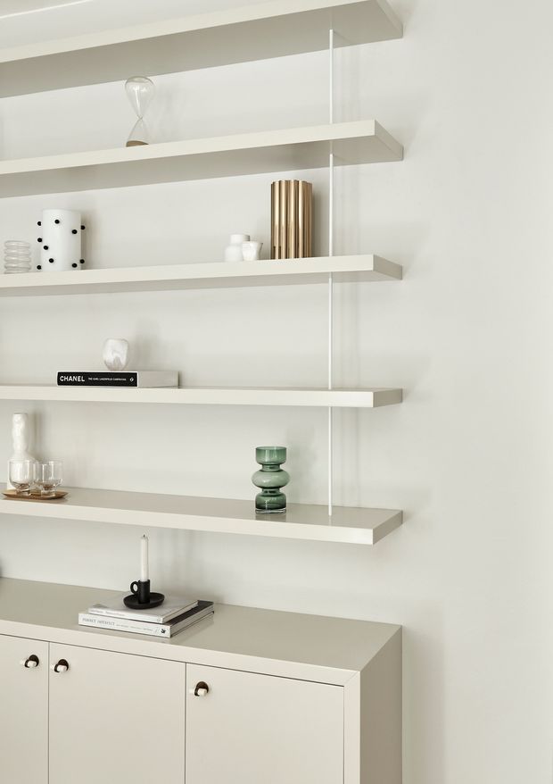 A minimalist interior vignette featuring a pale built-in shelving system with several long floating shelves above a low cream-coloured sideboard. The shelves display restrained decorative objects, books, candles, glassware and a sculptural brass-toned vase. The palette is soft white, cream and light grey with subtle dark and green accents, creating a calm, refined, highly ordered composition.