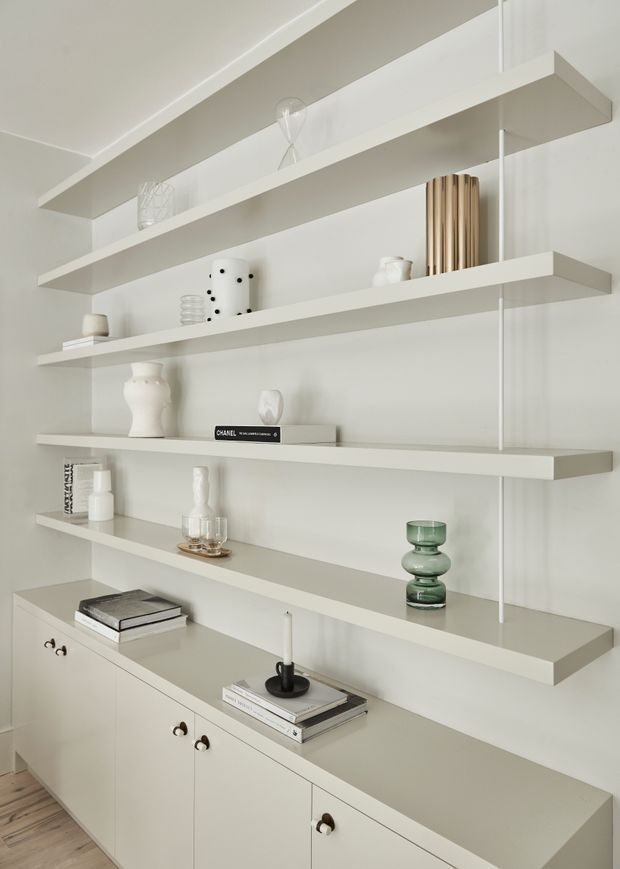 A bright, minimalist built-in display and storage wall with multiple long white floating shelves above a low run of closed cabinetry. The shelves are styled with neutral ceramics, books, glassware, and a few muted decorative objects, while the cabinetry has small dark metal or timber knobs. Light timber flooring and pale warm-white finishes create a clean, understated interior.