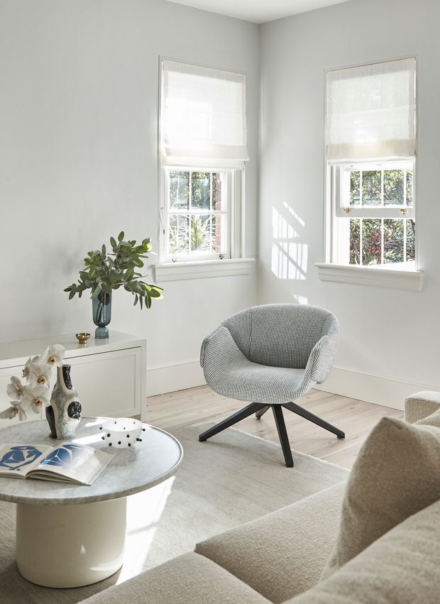 Bright, refined living room with pale walls, light timber flooring, two windows with white Roman blinds, and abundant natural light. The room features a sculptural patterned lounge chair, a neutral sofa, a round marble-topped coffee table, a white built-in cabinet, a pale rug, and understated decorative objects.