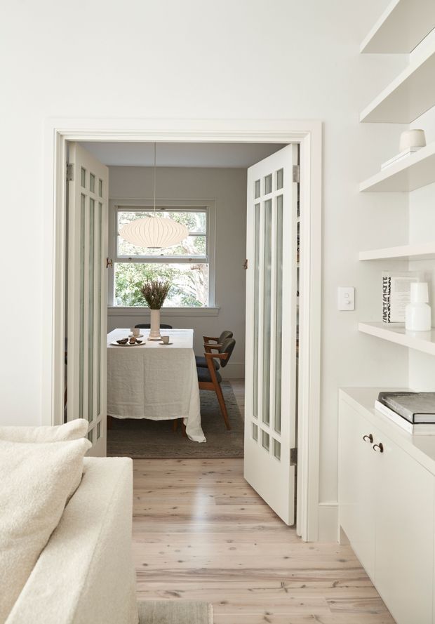 Bright, refined interior view from a living area through glazed double doors into a dining room. The space features pale timber flooring, white built-in shelving and cabinetry, classic framed doors, a linen-covered dining table, timber dining chairs, and a sculptural pendant light. The palette is soft and neutral with abundant natural light.