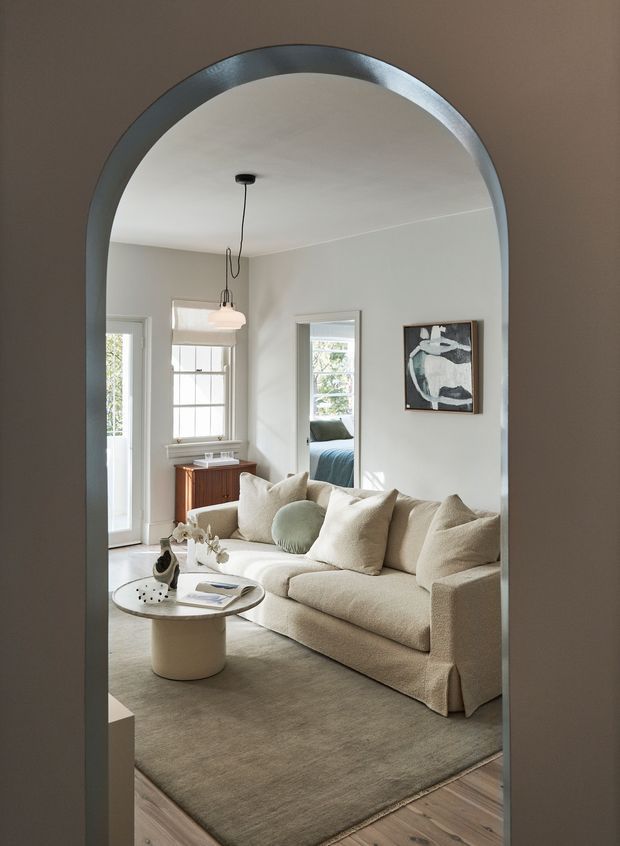 A softly lit, refined living room viewed through a large arched interior opening. The room features a neutral upholstered sofa with layered cushions, a round coffee table, a low timber sideboard, pale walls, framed artwork, pendant lighting, and timber flooring. A bedroom is visible through the internal doorway beyond the living area.