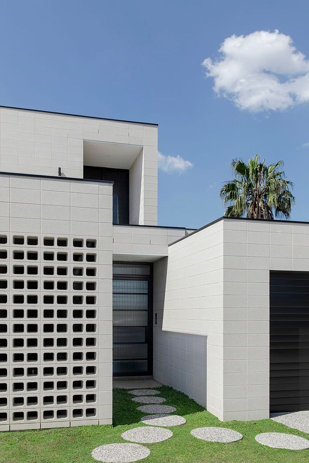 Exterior view of a contemporary multi-level residence with crisp white modular masonry cladding, flat rooflines, recessed entry, dark glazing and garage door, and a perforated breeze-block screen. A stepping-stone path crosses a lawn toward the entry, with a palm visible behind the building under a clear blue sky.