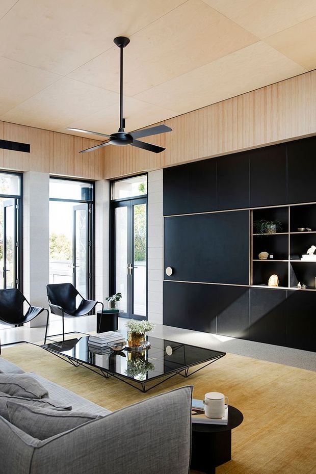 A bright, contemporary living room with a large grey sectional sofa, black leather lounge chairs, a geometric glass coffee table, and a dramatic full-height matte-black built-in media and display wall. Light timber wall panelling and ceiling panels contrast with the black joinery, while tall glazed doors bring in abundant daylight. A large black ceiling fan is centrally suspended above the seating area.