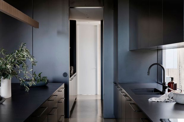 A dark, sophisticated galley kitchen with matte charcoal cabinetry and benchtops on both sides, integrated drawer storage, a deep undermount sink with a tall black gooseneck tap, and a narrow central passage leading to a bright white hallway. A timber open shelf and potted greenery add warmth and contrast. The space is compact, linear, and carefully detailed with a restrained contemporary aesthetic.
