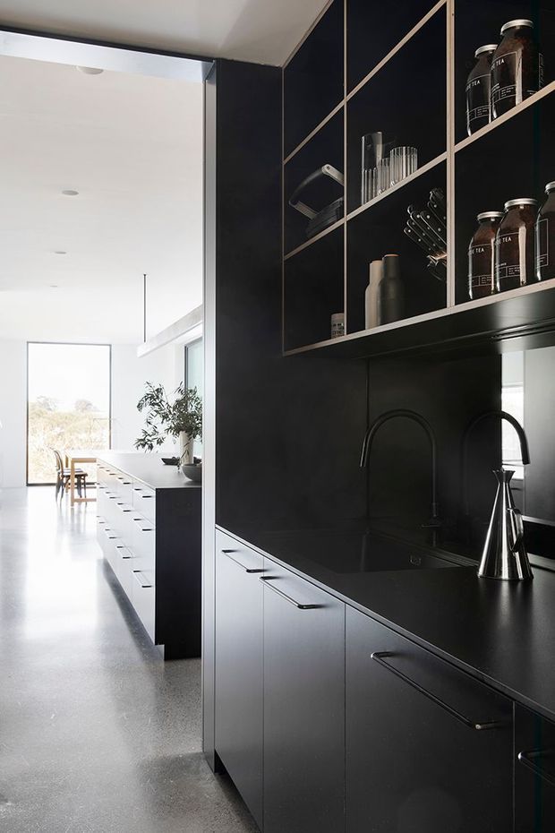 A sleek, predominantly black contemporary kitchen with extensive matte dark cabinetry, a dark benchtop and integrated open shelving. Black arched tapware, a sink, metallic kettle and neatly arranged tea jars and glassware create a refined functional composition. The kitchen opens to a bright dining area with white walls, full-height glazing and a long island, while a pale polished concrete or terrazzo-like floor extends through the space.