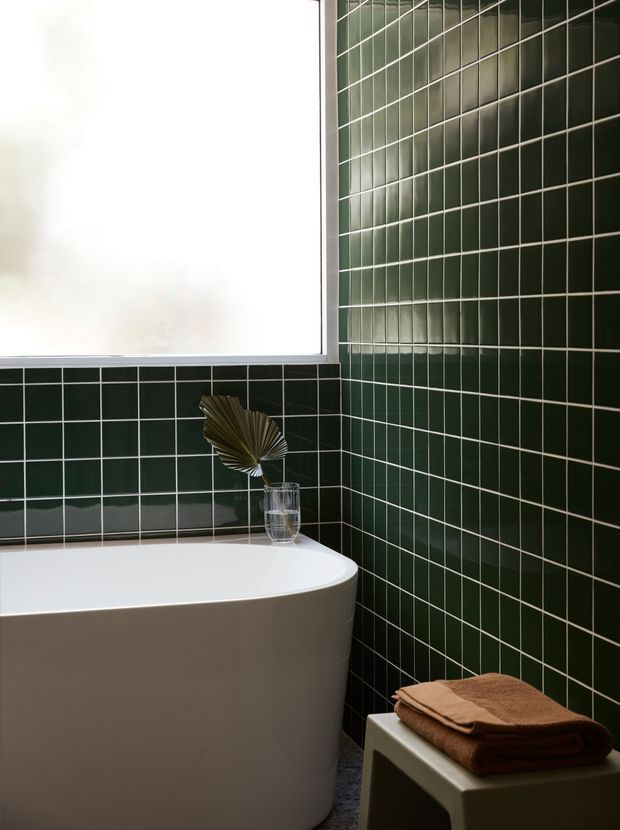 A compact, refined bathroom featuring a freestanding white bathtub set against glossy dark green square wall tiles with contrasting light grout. A frosted window provides soft natural light, while a small stool or side table holds neatly folded brown towels and a simple decorative arrangement.