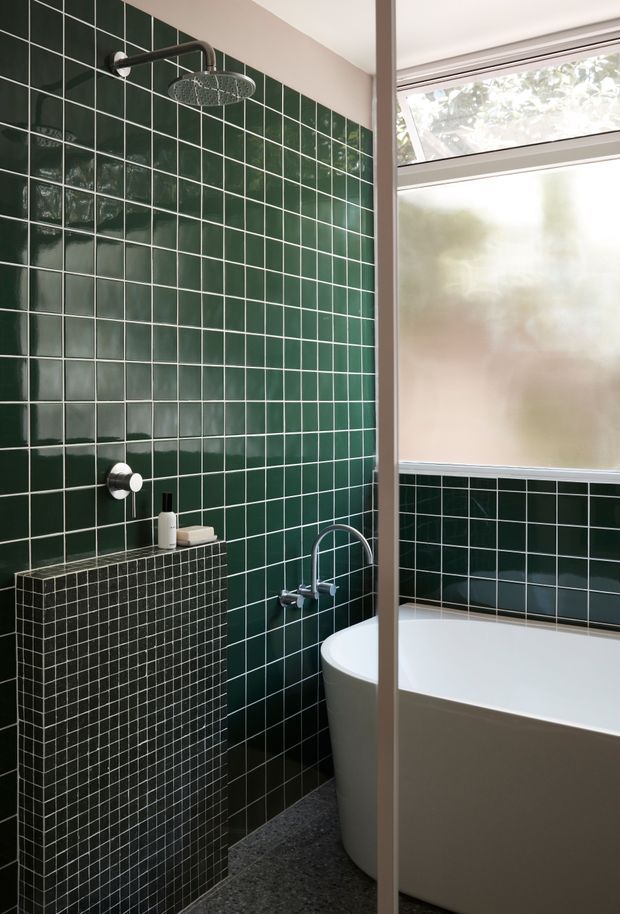 A compact contemporary bathroom featuring a deep forest-green glossy tiled shower wall, a matching dark mosaic-tiled half-height ledge, brushed metal shower fittings, a freestanding white bathtub, and a frosted window providing filtered natural light. The dark tiled surfaces contrast with the white sanitaryware and neutral trim.