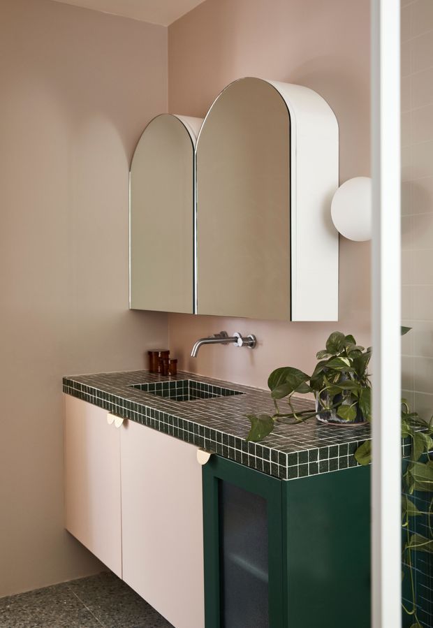 A compact, highly styled bathroom vanity with a muted blush-pink wall, pale pink cabinetry, dark green cabinetry accents, and a dark green small-format tiled benchtop with integrated tiled basin. Two tall arched mirrored cabinet doors hang above a wall-mounted chrome tap, accompanied by a spherical wall light and a trailing potted plant. The room has a warm, playful contemporary character and a terrazzo-like dark floor.