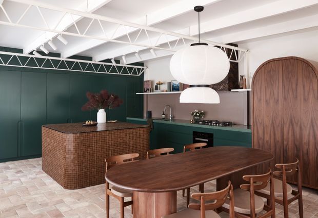 A sophisticated open-plan kitchen and dining space with deep green cabinetry, a rounded brown mosaic-tile island, dark timber dining table and chairs, and a dramatic arched timber door. White exposed roof trusses, track lighting, and large sculptural pendant lights contrast with the rich timber and green finishes.